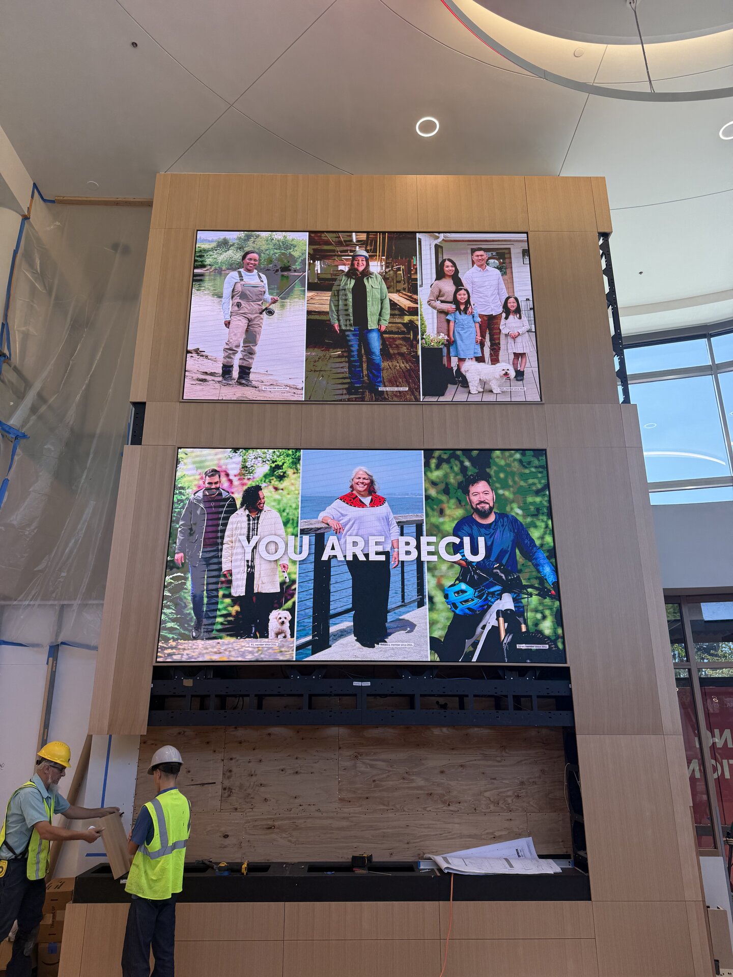 Crew installing a tall wood feature wall with two large digital displays reading You Are BECU
